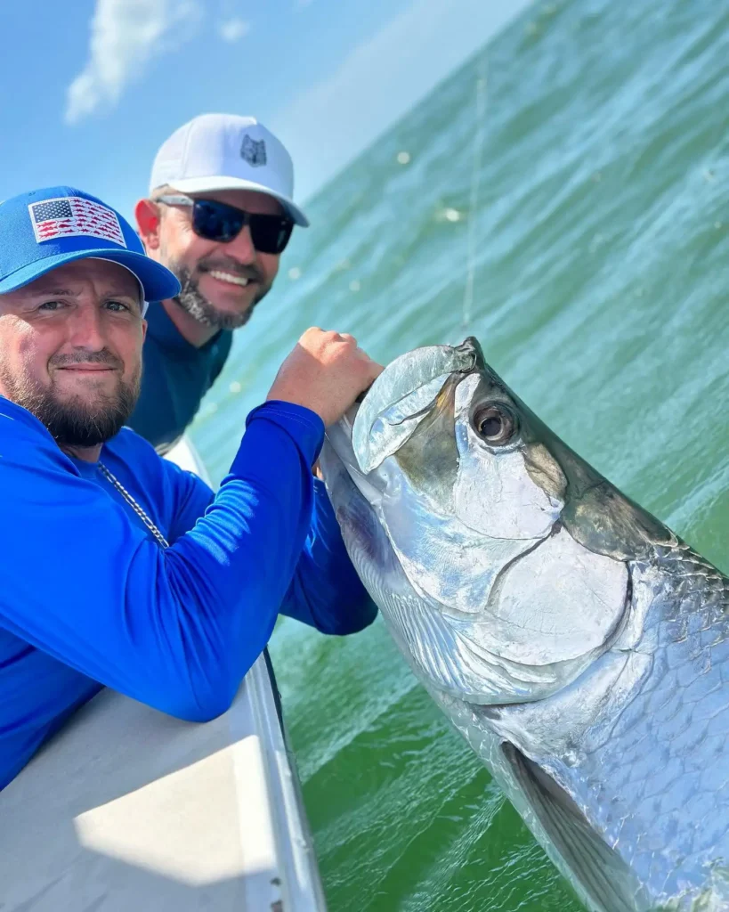 Two anglers holding a trophy tarpon beside a charter boat in Sarasota, Florida.