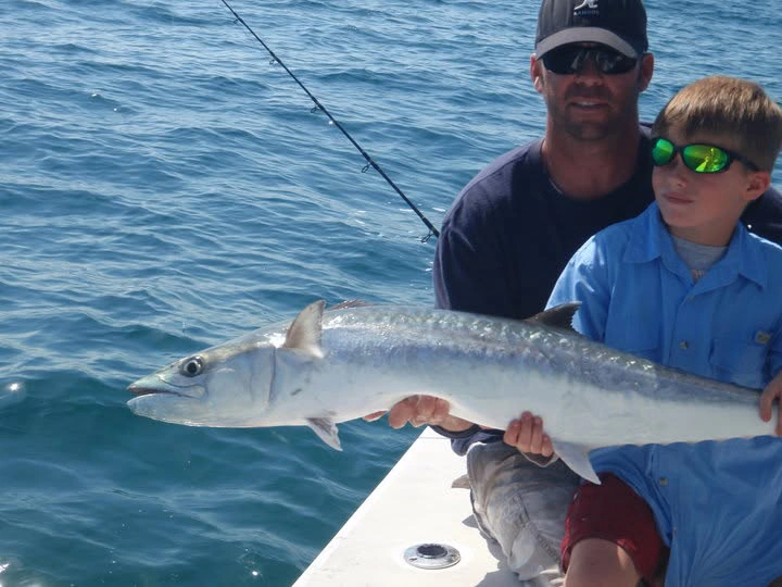 Young angler holding a large king mackerel aboard a Sarasota fishing charter boat with an adult beside him on calm Gulf waters.
